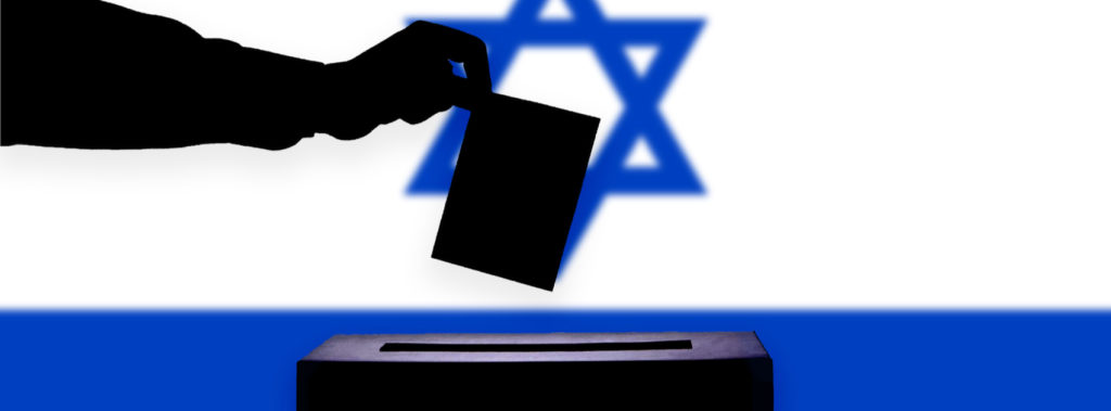Hand dropping election ballot into box in front of Israeli flag.