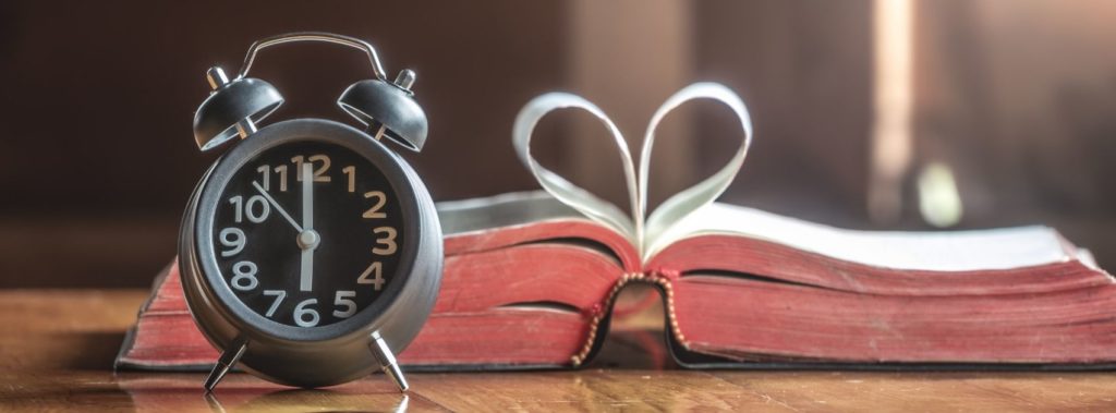 Clock next to open Bible with pages shaped like heart.