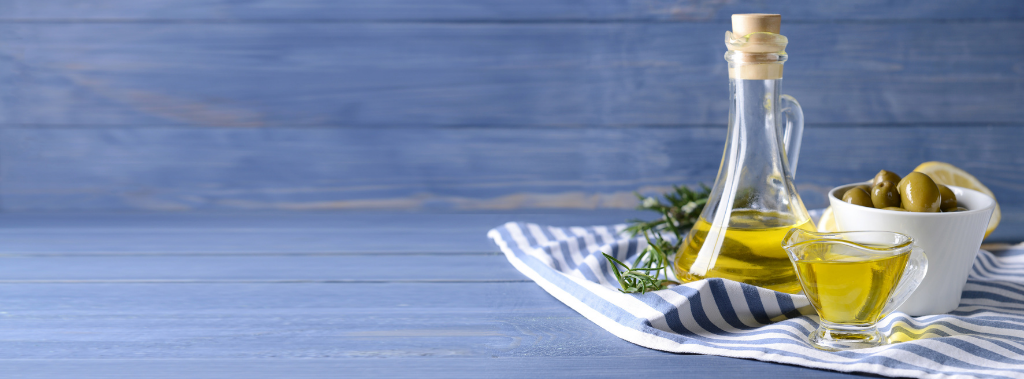 Glass bottle of olive oil with bowl of olives sitting on a blue striped towel.