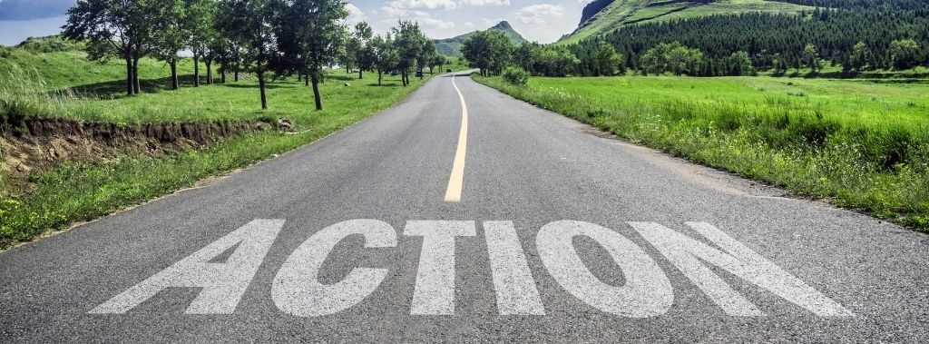 Country road with the word action written on it surrounded by lush greenery and a blue sky.