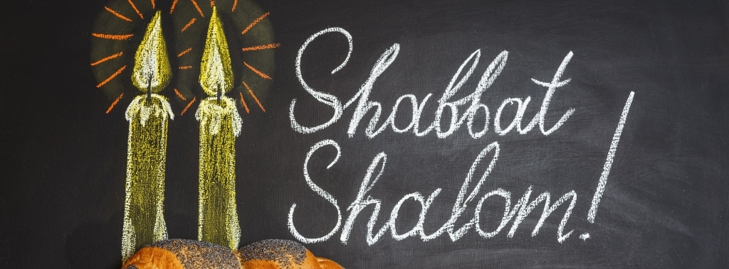 Words shabbat shalom and candles painted on chalkboard next to challah bread with poppy seeds.