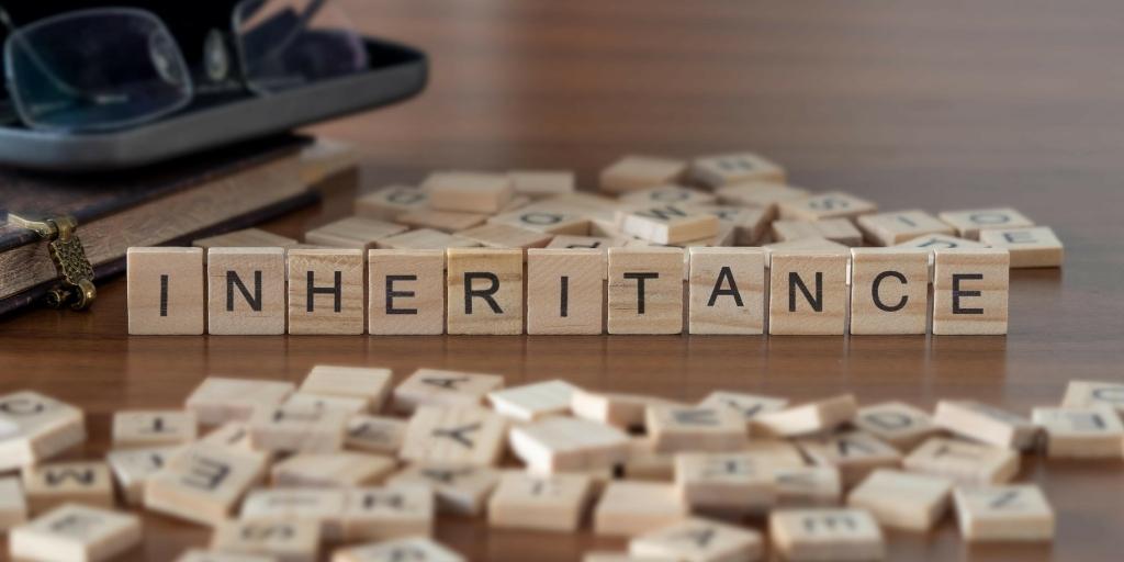 The word inheritance spelled out in wooden letter tiles on a table next to a book and glasses.